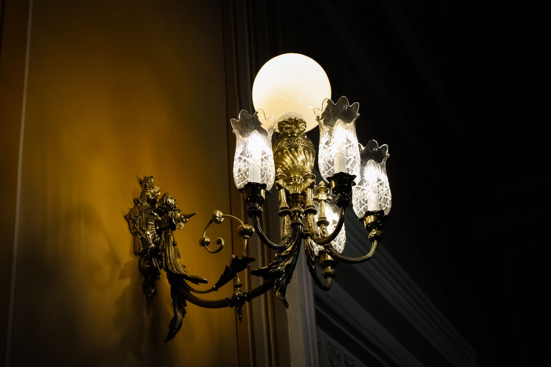 A beautifully ornate antique brass wall sconce lit in a dimly lit room, casting a warm glow.